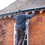 Man on a ladder fixing fitting a rain gutter on the eaves of a UK house